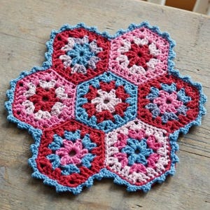 May include: A handmade, colorful crochet coaster or trivet. The hexagonal design features a floral pattern in shades of red, pink, and blue, with a scalloped blue border. The coaster is placed on a wooden surface.