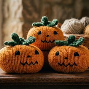 May include: Three round, orange knitted pumpkins with green stems and embroidered black facial features, including two eyes and a jagged mouth. The pumpkins are arranged in a cluster, showcasing a textured yarn construction and a soft, rounded shape.