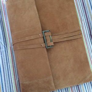 Puede incluir: Un diario encuadernado en cuero marrón con cierre de hebilla metálica. El diario está cerrado y colocado plano sobre una superficie de tela a rayas.