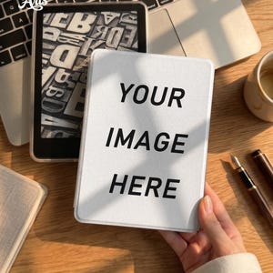 May include: A white e-reader case featuring the black text YOUR IMAGE HERE in a bold sans-serif font. The case is held against a wooden desk surface next to a laptop, a second e-reader displaying a black and white letter pattern, and a fountain pen.