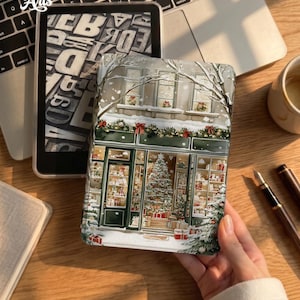 May include: A rectangular e-reader case featuring a festive winter scene of a storefront decorated with Christmas garlands, red bows, and a large decorated tree inside. The exterior shows snow-covered windows and a snowy sidewalk with gift boxes.