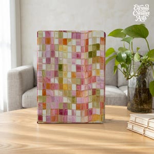 Könnte beinhalten: Ein rechteckiges Notizbuch mit einem Gittermuster aus kleinen Quadraten in Rosa-, Orange-, Gelb- und Olivgrüntönen. Der Einband hat eine glänzende, leicht verzerrte Oberfläche, die einen lebendigen, mehrfarbigen Mosaikeffekt erzeugt.