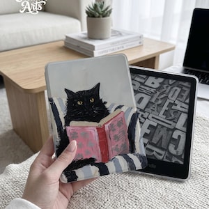 May include: A hand-painted Kindle case featuring a black cat reading a book with pink covers. The cat is seated on a striped cushion. The case is next to a Kindle device. The text "Eternal Creative Arts" is at the top.