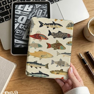 Könnte beinhalten: Eine beige Kindle-Hülle mit einem bunten Fischmuster. Die Hülle wird in der Nähe eines Laptops und einer Kaffeetasse gehalten. Sie zeigt verschiedene Fischillustrationen in Rot-, Blau-, Gelb- und Schwarztönen. Der Text "Eternal Creative Arts" ist sichtbar.