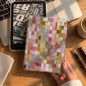 May include: A rectangular e-reader case featuring a mosaic pattern of small, glossy squares in shades of pink, orange, yellow, white, and olive green. The case has a smooth, reflective surface and is positioned next to a black e-reader device.