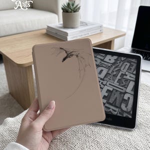 Può includere: Una custodia Kindle beige con un'illustrazione di un drago nero. La custodia è mostrata accanto a un e-reader Kindle che mostra del testo. L'immagine mostra anche il testo "Eternal Creative Arts" in corsivo.