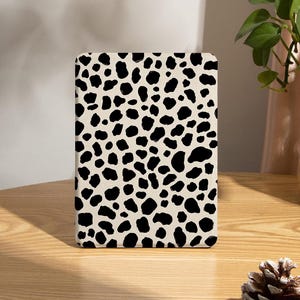 May include: A rectangular case with a cream-colored background and black spots, resembling a dalmatian pattern. The case is placed on a wooden table, with a plant in a glass vase and a pinecone in the background.