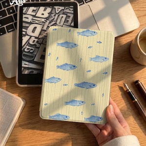 May include: A light yellow e-reader case featuring a pattern of blue fish and vertical lines. The case has a smooth texture and covers the device, which displays a black and white typographic design. The case is held in a hand over a wooden desk.