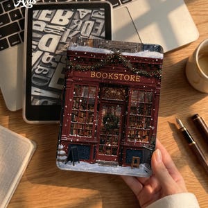 May include: A rectangular e-reader case featuring a detailed illustration of a dark red bookstore storefront covered in snow. The facade includes large glass windows, a central door with a wreath, and the word BOOKSTORE in gold lettering.