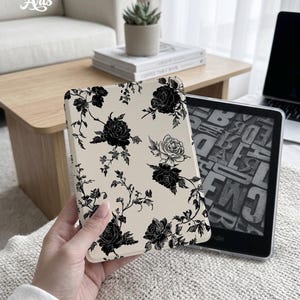 May include: A cream-coloured e-reader case with a black rose floral pattern. The case is held next to a Kindle e-reader. The text "Eternal Creative Arts" is visible in the upper left corner.