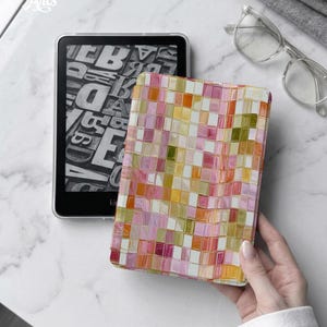 May include: An e-reader device featuring a black screen with a white typographic letter pattern. The device is encased in a protective cover displaying a vibrant mosaic of small, square tiles in shades of pink, orange, yellow, and olive green.