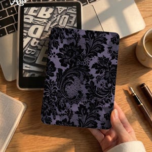 May include: A rectangular e-reader cover featuring a black lace pattern printed over a light purple background. The cover displays intricate floral and scrollwork motifs throughout the design, providing a detailed texture that covers the entire surface.