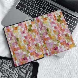 May include: A protective e-reader cover featuring a vibrant, multi-colored mosaic tile pattern in shades of pink, orange, yellow, and olive green. The cover is open, displaying a grid of small, glossy squares, and is positioned atop a silver laptop.