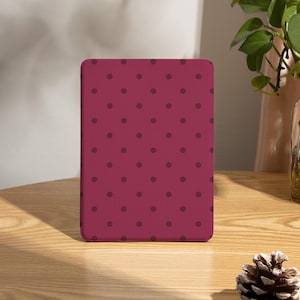 May include: A rectangular, burgundy-colored case with a repeating pattern of dark purple polka dots. The case is standing upright on a wooden table. A pinecone and a vase with greenery are also on the table.