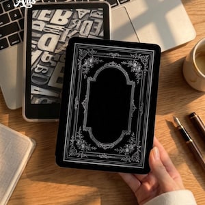 May include: A black e-reader skin featuring a white ornate rectangular frame with floral corner accents. The design includes a central blank space surrounded by intricate filigree patterns. The skin is applied to a rectangular device held in a hand.