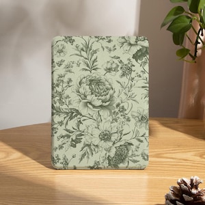 May include: A rectangular, pale green case with a floral design of peonies and other flowers in green and grey. The case is on a wooden table. A pine cone and a vase with greenery are also visible. The case is approximately 15 cm in height.
