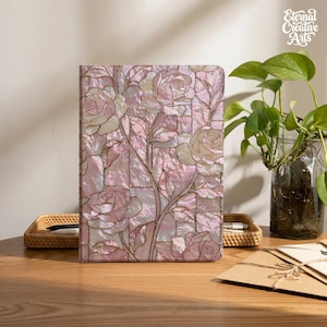 Könnte beinhalten: Ein rechteckiges Notizbuch mit einem Einband, der ein Mosaikmuster aus rosa Rosen und Blättern zeigt. Das Design verwendet schillernde, perlmuttartige Materialsegmente, die mit dünnen goldenen Linien umrandet sind und einen Buntglaseffekt erzeugen.