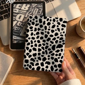 Op de afbeelding: Een rechthoekige e-reader hoes met een zwart-wit koeienvlekkenpatroon. De hoes heeft een glad, stevig oppervlak met onregelmatige zwarte vlekken verspreid over een witte achtergrond, wat zorgt voor een consistent ontwerp op de voorkant.