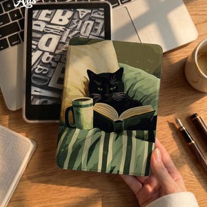 May include: A rectangular e-reader case featuring a graphic illustration of a black cat lying on a striped pillow, reading an open book next to a green mug. The case displays a cozy scene with muted green and cream tones, providing a protective cover.