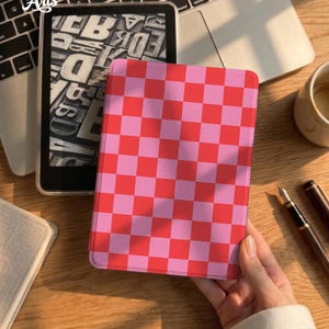 May include: A rectangular e-reader case featuring a vibrant red and light pink checkered pattern. The case is shown in a closed position, displaying a uniform grid of alternating colored squares across its entire front surface.