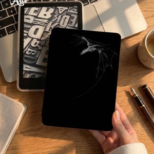May include: A black e-reader cover featuring a white line art illustration of a dragon in flight. The dragon is depicted in profile with wings extended against a solid black background. The cover is held in a hand, showcasing the detailed white graphic.