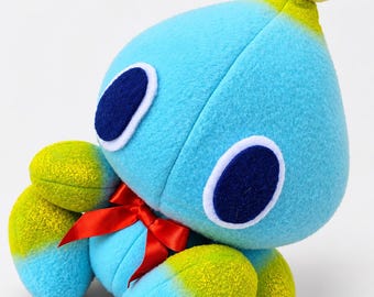 Puede incluir: Un peluche con forma de gota de tela azul claro, con una bola amarilla en la parte superior, ojos ovalados azul oscuro con bordes blancos y extremidades con puntas amarillas. Un pequeño lazo rojo brillante está atado al cuello.