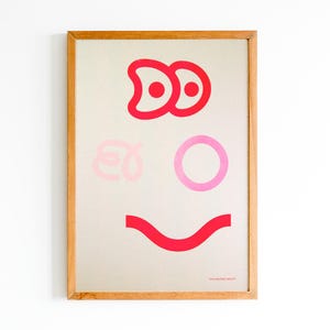 Pode incluir: Uma impressão de arte emoldurada com um design de rosto caprichoso. A obra de arte usa formas vermelhas e rosa para criar olhos, um nariz e uma boca sorrindo em um fundo neutro. A moldura é feita de madeira marrom clara.