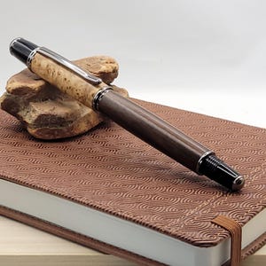 May include: A handcrafted wooden pen with a light-colored burl wood top and a dark brown wooden barrel. The pen features silver-colored metal accents and a black tip. It rests on a brown leather-bound journal with a patterned cover.