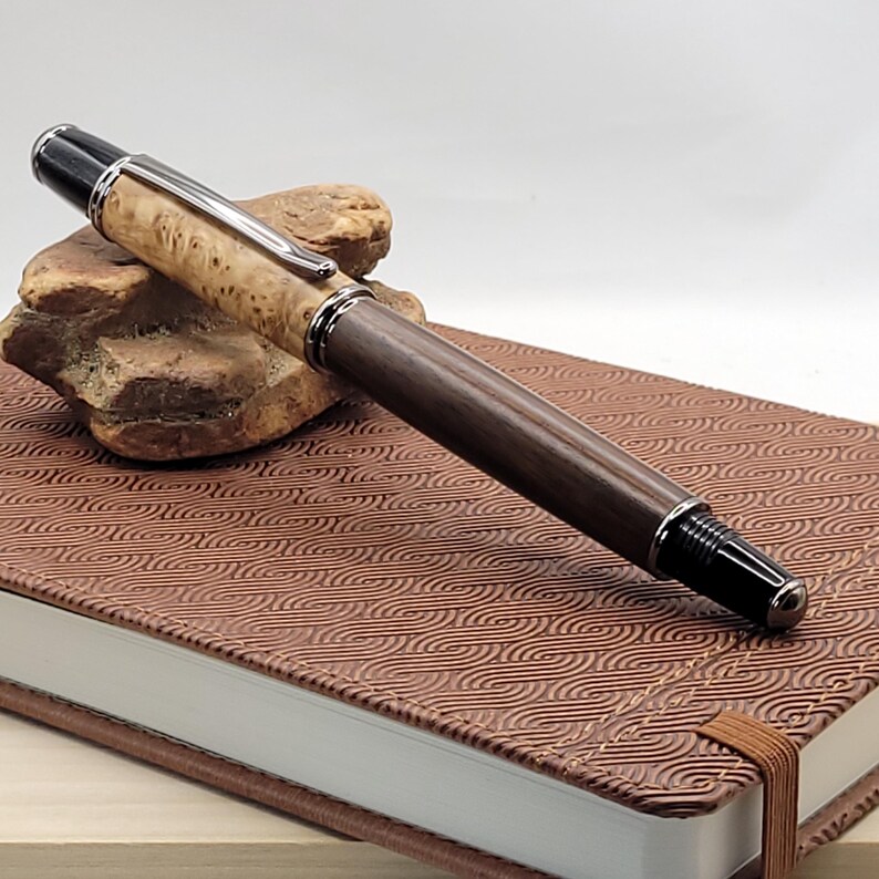 May include: A handcrafted wooden pen with a light-colored burl wood top and a dark brown wooden barrel. The pen features silver-colored metal accents and a black tip. It rests on a brown leather-bound journal with a patterned cover.