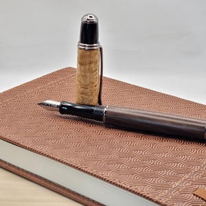 May include: A wooden fountain pen with a silver-colored cap and a black grip, resting on a brown leather-bound journal. The pen's body is a dark wood, and the cap is off to the side. The journal has a repeating wave pattern.