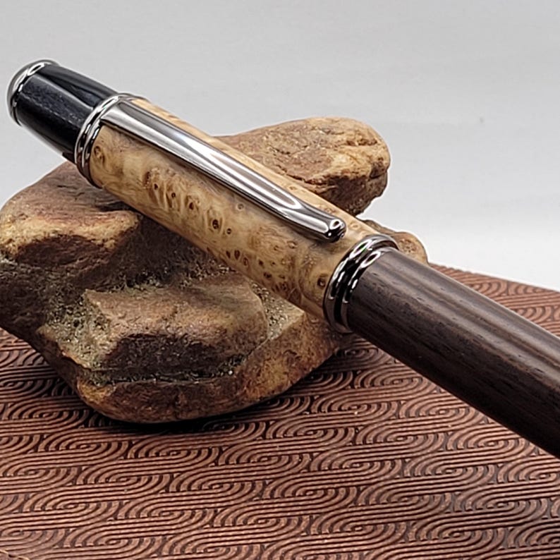 May include: A handcrafted pen with a light-colored, burl wood barrel and a dark wood lower barrel. The pen features silver-colored metal accents and a black top. The pen is displayed on a brown, textured surface.