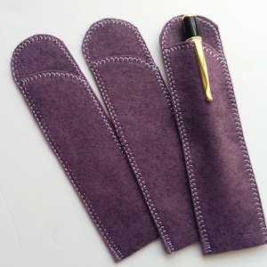 May include: Three plum-colored felt pen cases with white stitching. One case holds a gold and black pen. The cases are rectangular with rounded tops and are designed to protect pens.