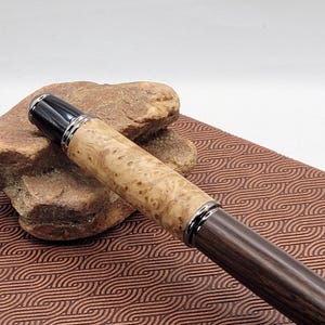 May include: A handcrafted pen with a light-colored, patterned wood barrel and a dark wood lower barrel. The pen features silver-colored metal accents and a black cap. The pen is displayed on a brown surface.