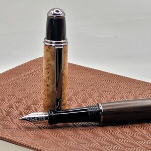May include: A fountain pen with a wooden barrel and a black cap is displayed. The pen's cap is detached, revealing the nib. The pen rests on a brown patterned surface. The pen's barrel has a light brown wood grain.