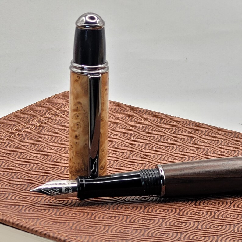 May include: A fountain pen with a wooden barrel and a black cap is displayed. The pen's cap is detached, revealing the nib. The pen rests on a brown patterned surface. The pen's barrel has a light brown wood grain.