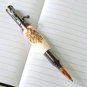 May include: A bolt-action pen featuring a textured, cream-colored antler barrel with a dark wood accent ring. The pen has a gunmetal finish on the cap and tip, with a copper-colored bullet-shaped point and a clip shaped like a rifle bolt.