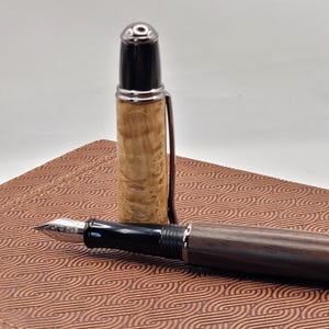 May include: A fountain pen with a wooden barrel and cap, and a silver-colored clip. The pen's nib is visible, and the cap is removed, revealing the pen's ink cartridge. The pen rests on a patterned, brown surface.