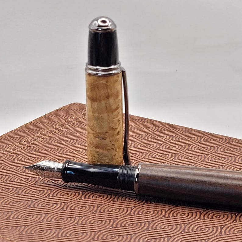 May include: A fountain pen with a wooden barrel and cap, and a silver-colored clip. The pen's nib is visible, and the cap is removed, revealing the pen's ink cartridge. The pen rests on a patterned, brown surface.