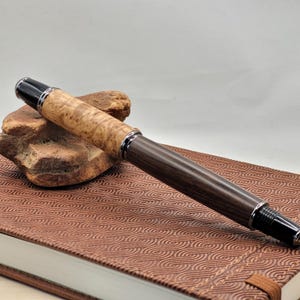 May include: A handcrafted fountain pen with a light-colored burl wood barrel and a dark wood grip. The pen features silver-colored metal accents and a black cap. It rests on a brown leather-bound notebook.