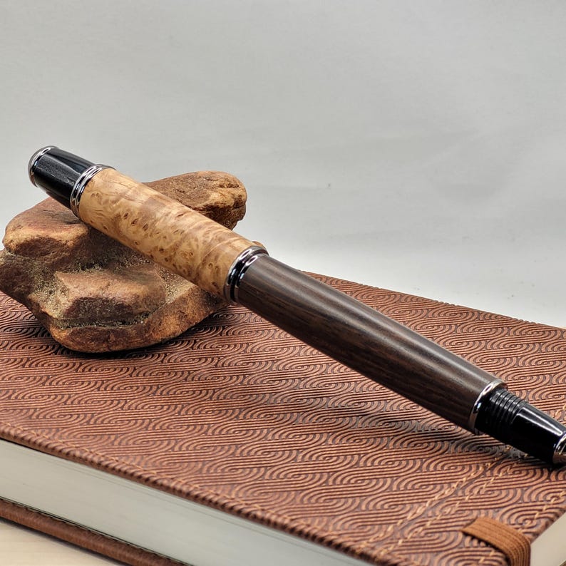 May include: A handcrafted fountain pen with a light-colored burl wood barrel and a dark wood grip. The pen features silver-colored metal accents and a black cap. It rests on a brown leather-bound notebook.