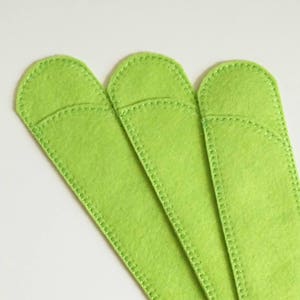 May include: Three lime green felt pen sleeves. Each sleeve is rectangular with a rounded top and a stitched border. The sleeves are arranged diagonally against a white background.