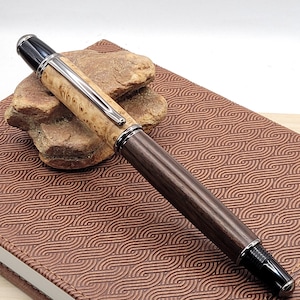 May include: A handcrafted pen with a light-colored, textured barrel and a dark wood lower barrel. The pen features silver-colored metal accents and a black tip and top. The pen rests on a brown notebook with a wave pattern.