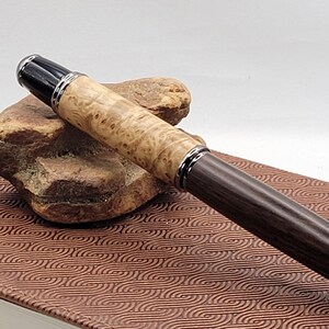 May include: A handcrafted pen with a light-colored, patterned wood barrel and a dark wood lower barrel. The pen features silver-colored metal accents and a black top. The pen is resting on a textured surface.