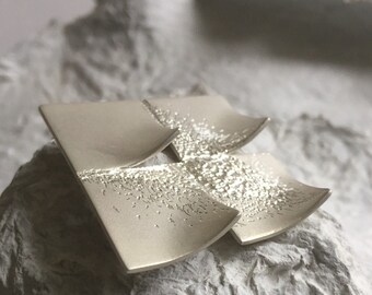 Puede incluir: Un broche de plata con un diseño geométrico texturizado. El broche está hecho de varias piezas superpuestas, creando un efecto de capas.
