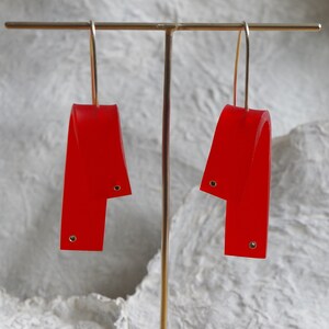May include: A pair of bright red, abstract earrings. Each earring features a curved, ribbon-like design with a small, circular accent near the bottom and top. The earrings hang from gold-colored hooks, displayed against a neutral background.