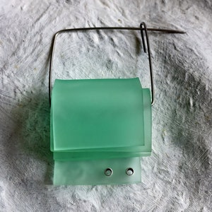 Könnte beinhalten: Eine grüne Glasbrosche mit einem Drahtrahmen und zwei Metallringen. Das Glas ist matt und hat einen geschichteten Effekt.