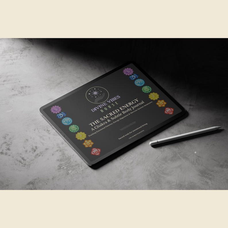 May include: A black tablet displaying the text "DIVINE VIBES HOUSE" and "THE SACRED ENERGY A Chakra & Subtle Body Journal". The tablet is surrounded by colorful chakra symbols and a white stylus rests beside it. The background is a textured gray surface.