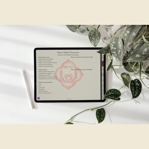 May include: A digital tablet displaying a "Root Chakra Overview" with text and a pink chakra symbol. A white stylus rests beside the tablet on a white surface, with green leafy plants in the background. The text includes "Balanced and Imbalanced Patterns."