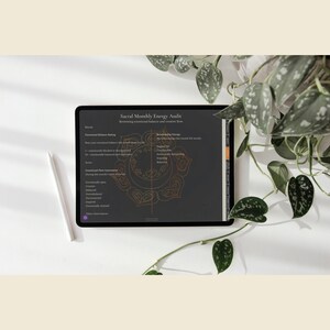 May include: A black tablet displaying a "Sacral Monthly Energy Audit" with text and a gold mandala design. A white stylus rests beside the tablet on a white surface, with green leafy plants in the background.