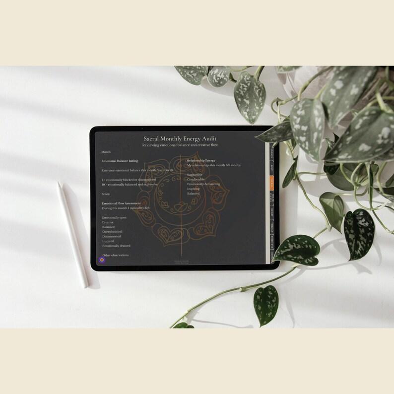 May include: A black tablet displaying a "Sacral Monthly Energy Audit" with text and a gold mandala design. A white stylus rests beside the tablet on a white surface, with green leafy plants in the background.